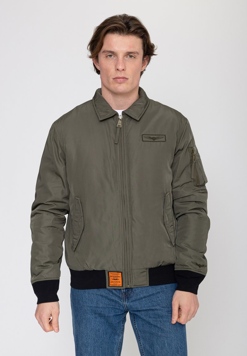 Uomo che indossa una giacca bomber verde oliva con zip frontale, toppa sul petto e polsini neri a coste, abbinata a jeans blu su sfondo semplice.