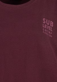 Burgundy cotton t-shirt featuring a round neckline and a printed logo in lighter burgundy on the left chest area. Smooth texture.