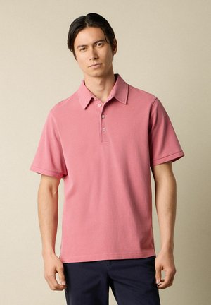 Giovane uomo con capelli scuri che indossa una polo rosa semplice a maniche corte e pantaloni scuri su uno sfondo beige.
