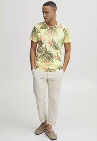 Camiseta amarilla de manga corta con estampado de hoja verde tropical y flor rosa, combinada con pantalones beige claros remangados y zapatos gris de slip-on.