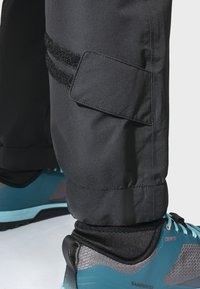 Schwarze Wanderhosen mit strukturiertem Finish und Klettverschlussdetails am Saum, kombiniert mit blauen und grauen Sportschuhen.