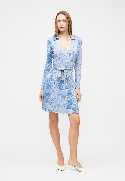 Γυναίκα με κυματιστά μαλλιά που φοράει μπλε φόρεμα με λουλουδάτο μοτίβο, μακριά μανίκια και δεμένη μέση, συνδυασμένο με λευκά μυτερά παπούτσια με χαμηλό τακούνι.