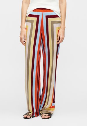 Luźne spodnie z pionowymi, wielokolorowymi paskami w kolorach czerwonym, niebieskim, beżowym, żółtym i brązowym, noszone z czarnymi sandałami i białym topem.