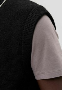 Zwarte gebreide vest met een textuur, gedragen over een licht taupe shirt met korte mouwen. De mouwen hebben een gladde afwerking en zichtbare stiksels.
