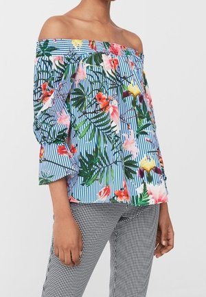 Femme portant un chemisier épaules dénudées à rayures bleues et blanches avec un imprimé floral tropical coloré, associé à un pantalon à carreaux noir et blanc.