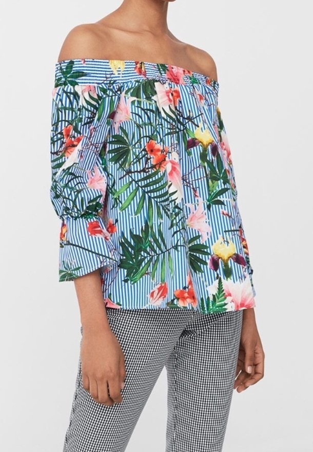 Femme portant un chemisier épaules dénudées à rayures bleues et blanches avec un imprimé floral tropical coloré, associé à un pantalon à carreaux noir et blanc.