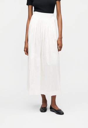 Žena oblečená v bielej plisovanej midi sukni s vysokým pásom, čiernom krátkom tričku a čiernych baletných balerínach, stojaca na jednoduchom pozadí.