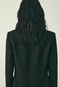 Čierny blejzer s hladkou textúrou a štruktúrovanými ramenami, s centrálnym švom pozdĺž zadnej časti a dlhými, rovnými rukávmi.