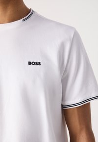 Bílý tričko s kulatým výstřihem vyrobené z bavlny, s malým černým logem "BOSS" na levém hrudníku a černými pruhovanými akcenty na límci.
