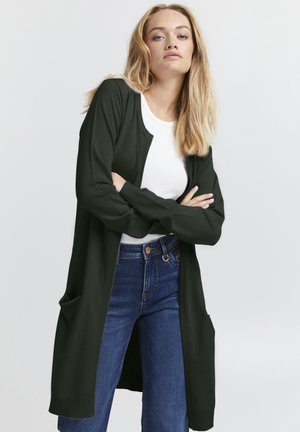 Cardigan verde scuro con maniche lunghe, apertura frontale e tasche laterali, abbinato a un top bianco e jeans blu. Texture liscia e design casual.