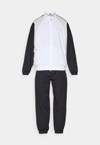 Tracksuit med en hvid lynlåsjakke med sorte og grå accenter og matchende sorte bukser med elastiske manchetter. Fremstillet af et glat stof.
