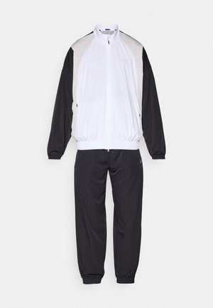 Fato de treino com um casaco branco com fecho éclair e detalhes em preto e cinza, e calças pretas combinando com punhos elásticos. Feito de um tecido suave.