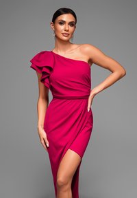 Robe fuchsia à une épaule avec des détails en volants, corsage ajusté, fente sur le côté et texture drapée, associée à des boucles d'oreilles étincelantes.