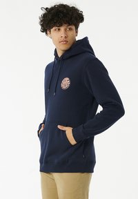 Marineblauwe hoodie met een kangoeroe-pocket, een trekkoordcapuchon en een ronde roze grafische patch op de borst. Zachte stof met geribde manchetten.