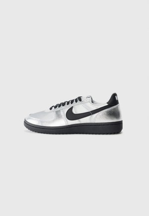 Baskets basses argentées avec logo swoosh noir, lacets noirs et semelle noire, vues de l'intérieur sur un fond gris.