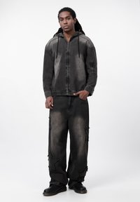 Zwarte denim hooded jack met een gedeeltelijk ritssluiting aan de voorkant, gecombineerd met brede zwarte jeans met stervormige uitsnijdingen en rafelranden.
