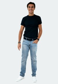 Schwarzes T-Shirt mit kurzen Ärmeln, hellblaue Slim-Fit-Jeans, weiße Sneakers und einen schwarzen Gürtel. Das Model steht mit den Händen in den Taschen vor einem hellen Hintergrund.