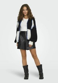 Cárdigan de punto en negro, blanco y azul con un patrón de tejido texturizado. Llevado con una blusa blanca y pantalones cortos de cuero negra. Botas negras hasta la rodilla.