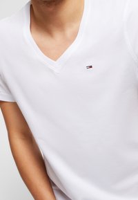 Bílý bavlněný tričko s výstřihem do V, které má na levé straně hrudníku malé vyšívané logo s červenými, bílými a modrými pruhy. Hladký povrch.