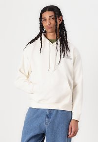 Cream hoodie with drawstring, front pocket, and embroidered "Obey Studios" logo. Worn with light blue denim jeans. Smooth texture.
