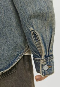 Denimjacka i blek blå färg, med knäppningar vid ärmsluten, skräddarsydd passform och klyvning på ärmen.