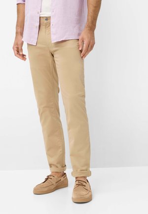 Mann trägt beige hochgekrempelte Chinos, braune Wildleder-Bootsschuhe, ein weißes Unterhemd und ein hellviolettes Hemd vor weißem Hintergrund.