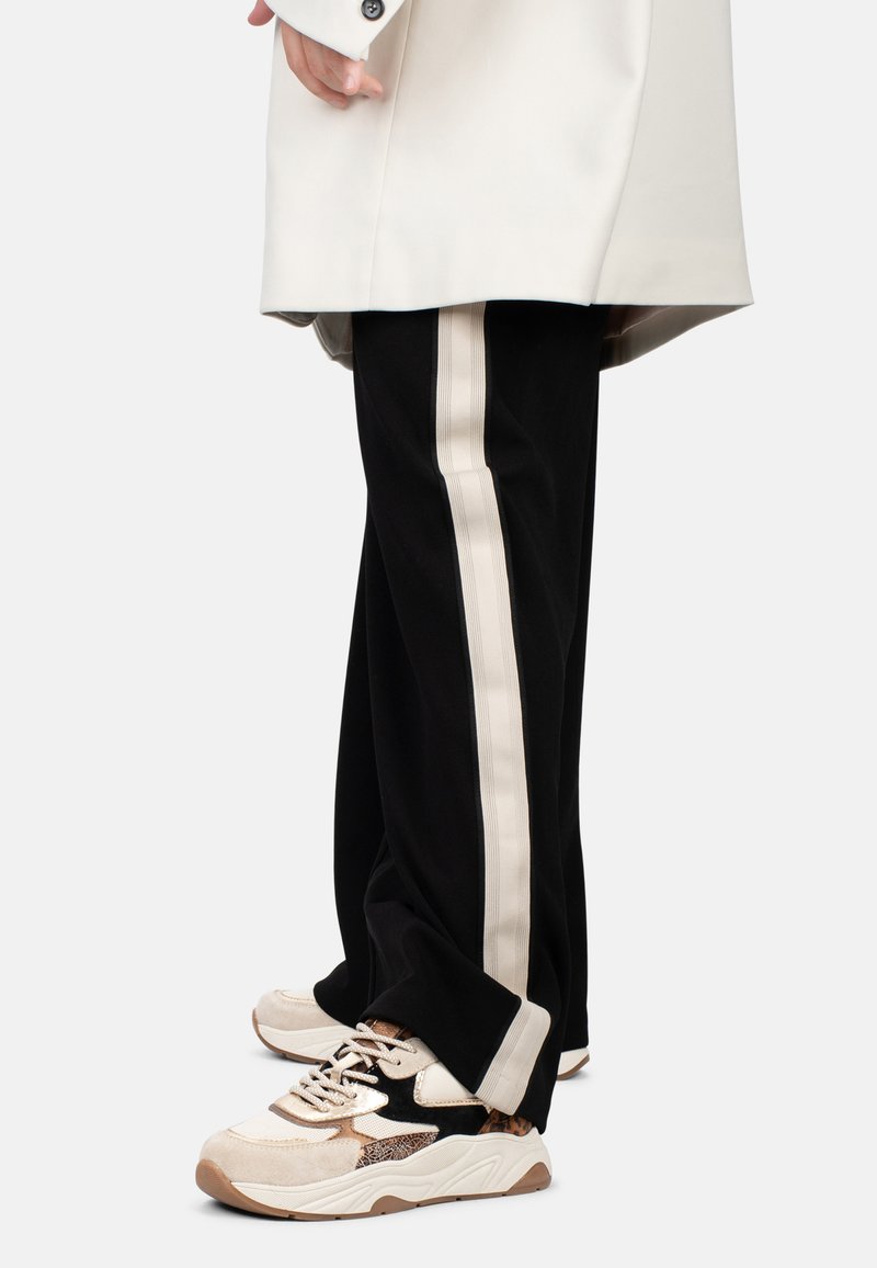 Pantaloni neri con una striscia beige sui lati, abbinati a sneaker crema e con motivo. I pantaloni hanno una vestibilità rilassata e un tessuto testurizzato.