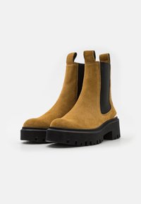 Stivali Chelsea in suede marrone con pannelli elastici neri, suole in gomma spessa e un motivo di battistrada dettagliato. Altezza alla caviglia con passanti per facilitare l'indosso.