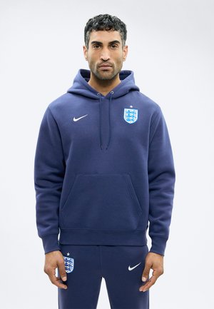 Mand iført marineblå England fodbold-hoodie og matchende bukser med Nike-logoer og England holds våbenskjolde, stående foran en ensfarvet baggrund.