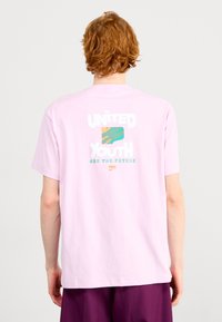 Osoba z czerwonymi włosami, ubrana w jasnoróżową koszulkę z napisem i grafiką "MŁODZIEŻ ZJEDNOCZONA TO PRZYSZŁOŚĆ" na plecach, w zestawieniu z fioletowymi spodniami.