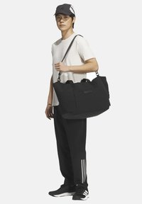 Borsa da palestra nera con manici superiori e tracolla, caratterizzata da una texture liscia e un design minimalista. Indossata da una modella in abbigliamento sportivo.