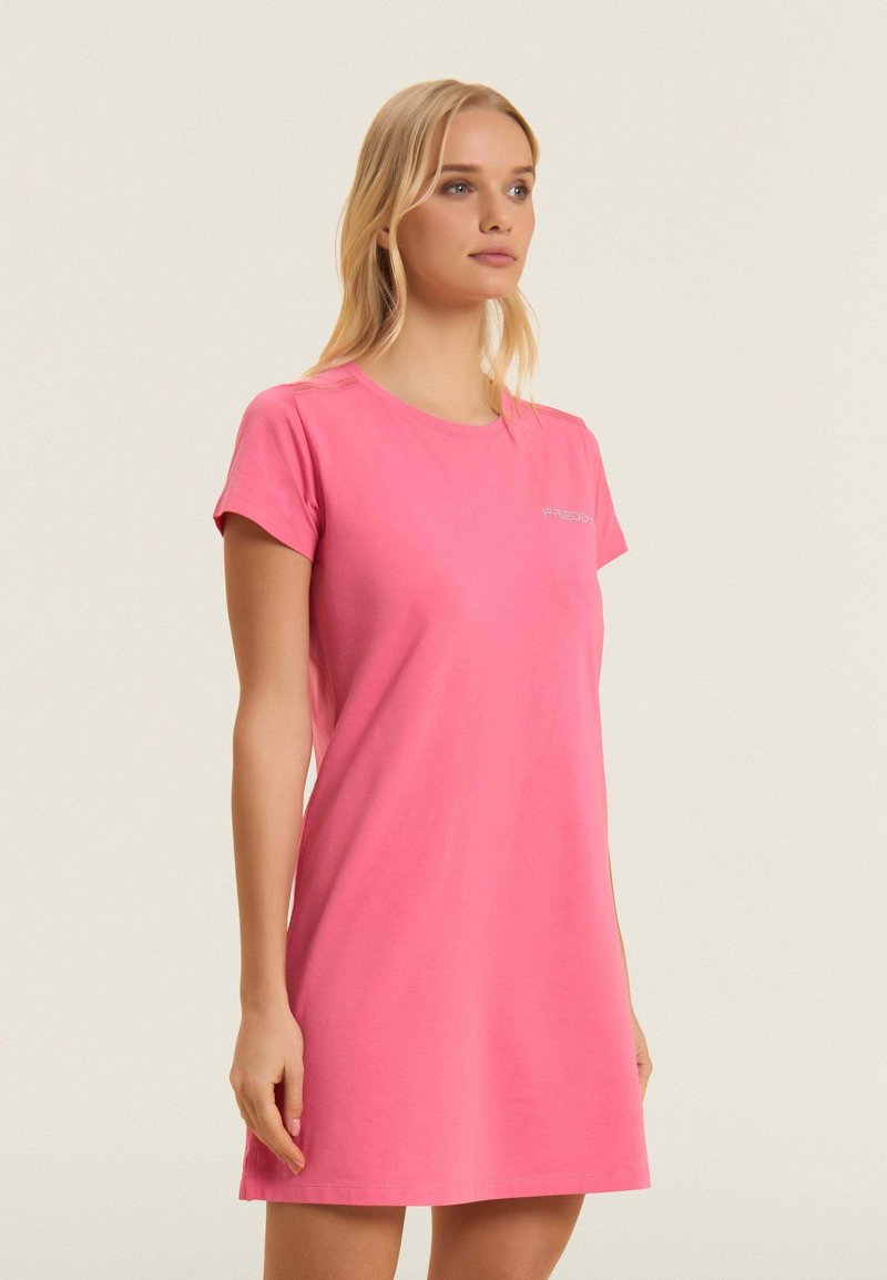 Femme portant une robe décontractée rose à manches courtes, debout avec les mains détendues le long du corps, regardant légèrement vers la droite sur un fond uni.