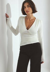 Femme aux cheveux bruns courts portant un haut blanc ajusté à manches longues en V et un pantalon noir, appuyée contre un mur blanc.