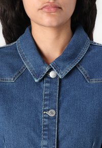 Casaco de ganga azul com gola, fecho de botão prateado e detalhes de costura visíveis. Apresenta um ajuste estruturado e um tecido texturizado.