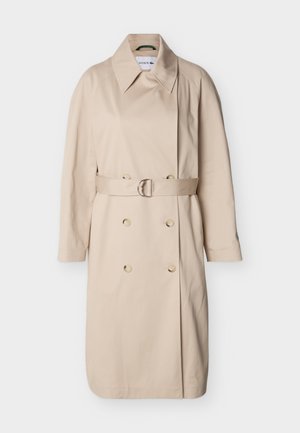 Gabardina beige cruzada con cuello ancho, mangas largas, cinturón con hebilla plateada en la cintura y detalles de botones en la parte delantera.