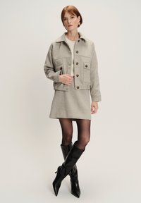 Giacca grigia in lana con chiusura a bottone, due tasche frontali e colletto, indossata sopra un maglione chiaro e una gonna grigia; abbinata a stivali neri al ginocchio.
