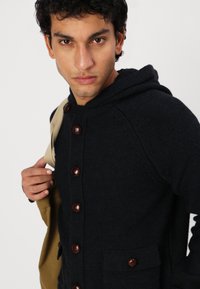 Pulover cu glugă de culoare bleumarin, cu tricot texturat, având butoane mari maro, două buzunare frontale și o croială lejeră. Modelul ține o geantă bej.