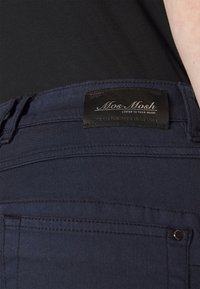 Pantalones de mezclilla azul oscuro con una etiqueta negra de Mos Mosh en la cintura. Incluye detalles en los bolsillos traseros y costuras cosidas.