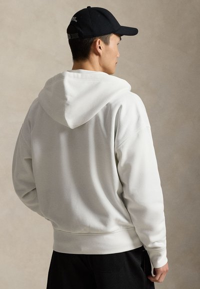 Sweat-shirt à capuche blanc de coupe décontractée, présentant une texture douce et des poignets côtelés, porté avec un pantalon de sport noir et une casquette sombre.