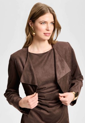 Femme aux cheveux châtain clair portant une veste en daim marron et une robe, des boucles d'oreilles créoles dorées et un large bracelet doré à son poignet.