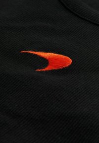 Svart stickad tyg med en orange broderad logotyp i en kurvad form, placerad på sidan. Den rika texturen ger djup åt designen.