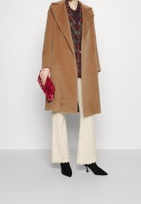 Bruine wollen jas met grote revers, over een geruite vest, gecombineerd met crème gekleurde flared broeken met textuur en zwarte puntige hakken. Houdt een rode clutch vast.