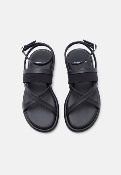 Μαύρα επίπεδα σανδάλια με διασταυρούμενους ιμάντες και αγκράφες στον αστράγαλο, σε λευκό φόντο, σχεδιασμένα για καθημερινή χρήση.