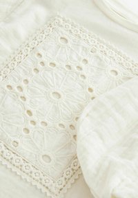 Tissu blanc présentant une délicate broderie florale avec des zones dégagées, bordé d'une dentelle complexe. Texture lisse avec un subtil éclat.