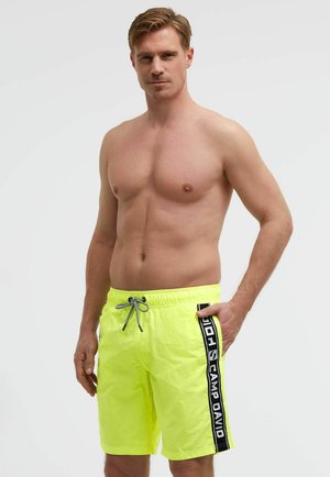 Oberkörperfrei stehender Mann in leuchtend gelben Badeshorts mit schwarzem und weißem "Camp David"-Seitenstreifen vor schlichtem Hintergrund.