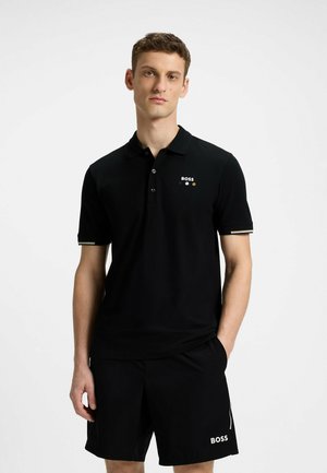 Black polo shirt with short sleeves, three buttons, and brand logo in white and gold accents. Worn with black shorts featuring a logo.