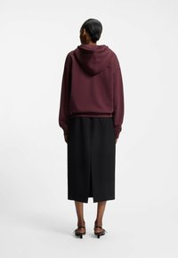 Felpa con cappuccio color marrone scuro dalla vestibilità rilassata, abbinata a una gonna nera al ginocchio con spacco posteriore e sandali neri con cinturini.