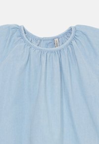Lyseblå, myk stoffbluse med samlet halsutskjæring og løs passform. Har et enkelt design og en etikett med merkevarenavn synlig.