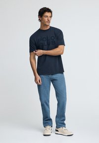 Mörkblå t-shirt med stor "POLO CLUB" text, rundad halsringning, tillsammans med ljusblå jeans och beige sneakers. Enkelt och avslappnat design.