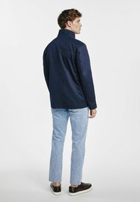 Granatowa kurtka z gładkiego materiału, z kołnierzem stojącym i długimi rękawami. W zestawieniu z jasnoniebieskimi dżinsami i ciemnymi trampkami.